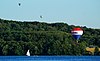 Three hot air balloons over a lake with a sailboat and forested shore