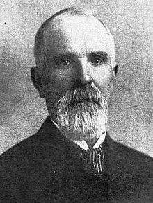 Black-and-white photograph of a man with thin hair and a beard in a suit and tie, with a stern expression