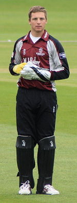 A young man with light brown hair is standing on some grass. He is wearing a burgundy, grey and black cricket uniform, large gloves and black trousers covered by black think pads on his lower legs.