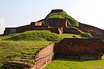 Ruins of a structure of red stone now resembling a small hill or mound.