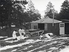 A shack surrounded by pine trees. There is snow on the ground. A man and a woman in white lab coats are pulling on a rope, which is attached to a small trolley on a wooden platform. On top of the trolley is a large cylindrical object.