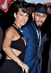 A woman wearing a black dress and a man wearing a suit look toward a camera