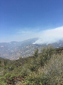 Thin gray smoke spreads out from an unseen fire burning on steep, scrubby mountainside across a valley from the picture's vantage point