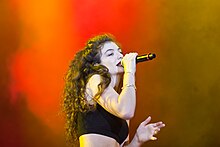 Lorde performing onstage against a background mixing shades of red and orange