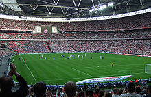 The interior of an empty stadium as viewed from its upper tier of seating. The seats are a vivid red and the pitch is a vivid green. The pale grey sky is visible through an opening in the ceiling above the pitch.