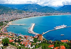 A colorful city with red roofs rising out from a curving harbor with blue water and cruise ship docked by a long pier.