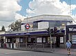 A grey building with a blue sign reading "EALING COMMON STATION" in white letters and two green trees in the background all under a blue sky