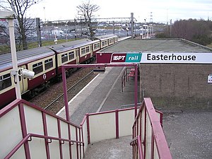 Two railway tracks with overhead catenary electrification pass through a railway station with a small, single-storey, flat-roofed station building and stairs presumably leading to a footbridge