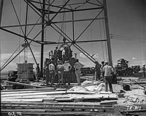 Men stand around a large oil-rig type structure. a large round object is being hoisted up.