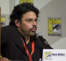 A bearded, dark-haired man leans into a microphone; he is sitting behind a San Diego Comic Con paper placard that declares his name "Kenneth Biller".