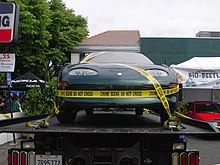 Front view of a dark green car, with yellow caution taping exclaiming, "CRIME SCENE DO NOT TOUCH". It is on the tray of a large truck.