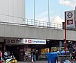 A number of people standing and sitting in front of a grey building with a white sign reading "Ealing Broadway" all under a blue sky with white clouds