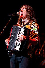 Una fotografía de "Weird Al" Yankovic, cantando a través de un micrófono y tocando una armónica.