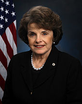 A woman wearing a suit and a necklace stands in front of a U.S. flag