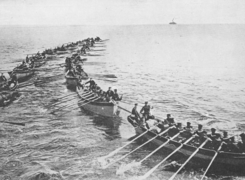 photo d'un groupe de chaloupes. Sur deux rangs, ces bateaux sont remplis de soldats en train de ramer.