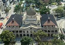 The grey dome and front gable of a granite neo-classical building, with a skyscraper in the background against a clear blue sky