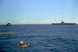 Photographie en couleur de la récupération en mer des astronautes d'Apollo 16. Le module de commande Casper flotte sur l'eau et des hommes se trouvent sur un bateau pneumatique à côté du module. L'hélicoptère de récupération, un Sikorsky SH-3 Sea King, les survole. Le porte-avions USS Ticonderoga est visible à l'arrière-plan.