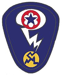 Oval shaped shoulder patch with a deep blue background. At the top is a red circle and blue star, the patch of the Army Service Forces. It is surrounded by a white oval, representing a mushroom cloud. Below it is a white lightening bolt cracking a yellow circle, representing an atom.