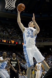 A basketball player wearing a white jersey lays taking a shot close to the rim.