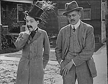 Capture d'écran montrant deux hommes en costume dans une rue. Chaplin porte un haut-de-forme, une redingote et la moustache tombante.