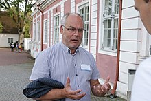 Frontale Farbfotografie von einem Mann, der zu einer Person am rechten Bildrand spricht. Mit seinen Händen gestikuliert er angespannt vor seiner Brust. Er hat lichte, graue Haare und trägt eine Brille und einen Dreitagebart. An seinem kurzärmeligen, blauen Hemd steckt rechts ein Namensschild und er hat sein dunkelblaues Jackett über seinen rechten Arm gelegt.