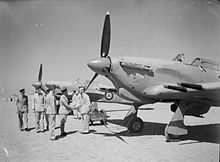 Photographie noir et blanc de deux avions au sol. Un groupe de militaire se déplacent devant et deux hommes se serrent la main.