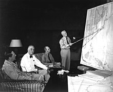 Three men are seated in lounge chairs. One is standing, holding a long stick and pointing to the location of Japan on a wall map of the Pacific.