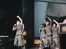 Image of a woman wearing military gear. She is blonde and is wearing a hat on her head. She's surrounded by people dressed in similar clothing.