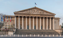 Frontale Farbfotografie eines klassizistischen Gebäudes mit einem breiten Säuleneingang und fünf grünen Glastüren. Über den Säulen steht in Gold „Assemblée Nationale“ und im Giebelfeld ist eine antike Szene um einen Thron. Auf dem Dach ist eine Frankreichflagge und links neben dem Gebäude wehen drei Frankreichflaggen und eine Europaflagge. An beiden Seitenmauern sind in schmalen Rechtecken antike Szenen. Links und rechts neben den Außenstufen sind Steinfiguren.