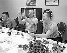 Photographie en noir et blanc Fred Haise, James Lovell, et Jack Swigert assis à une table pour un repas.