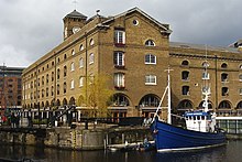 Converted dock warehouse in buff brick with a prominent parapet. A gap is visible in the parapet above the column of loading doorways, where a lucarne has been removed. The doorways also have hinged iron flaps, restrained by chains, as loading extensions.