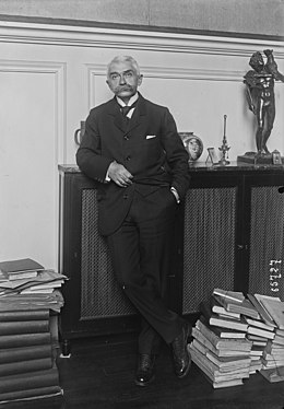 Portrait photographique de Pierre de Coubertin se tenant debout, main dans le poche, en costume, entre des piles de livres posées au sol.