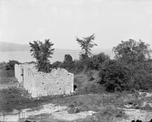 A black-and-white photograph. Ruined wall sections are visible in the center, with a tree growing out of the center of one of the structures. The lake is visible in the background, and hazy land is visible even further back.