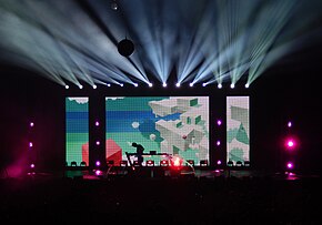 On a distant stage, many lights shine outward, and Porter Robinson is silhouetted against a screen depicting the sky and floating cuboidal structures.