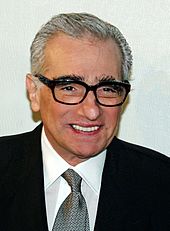 A man with grey hair in a dark suit, white shirt and grey tie, wearing glasses