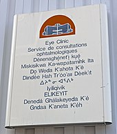 A metallic white sign on a gray background with a red and blue depiction of a building at the top. Below it is text in blue saying "Eye Clinic" in English, French and the other nine official territorial languages