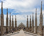 A stone rooftop, sloping down from a level centre to edges where intricately decorated stone spires rise, with statues atop, proceeding toward a rear wall with some more spires. Near the walls, people are gathered. Above is a blue sky largely covered with thin clouds