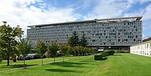 In Geneva, Switzerland, a large rectangular building stands beside a lush green lawn and a row of trees in the middle