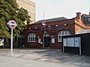 A red-bricked building with a blue sign reading "BOW ROAD STATION" in white letters and a tree in the foreground all under a blue sky with white clouds