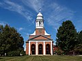 The front of a brick church with white trim. There are three arched openings, lined on each side with white pillar-like facings. The tower has a pillared section in which the church bell is visible, and it is topped by a cupola and a weathervane.