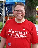 A man in a red T-shirt and glasses smiles while outdoors on a sunny day.