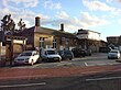 A brown-bricked building with three brown-bricked chimneys, some scaffolding on the side, and seven vehicles in the parking lot in front