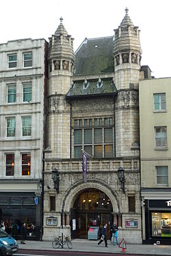 grey stone building with arched entrance and two flanking towers from the first floor with a steep-pitched grey roof with plain multi-storeyed buildings on either side
