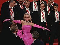 Monroe in Gentlemen Prefer Blondes. She is wearing a shocking pink dress with matching gloves and diamond jewellery, and is surrounded by men in tuxedos