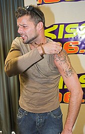 Ricky Martin olhando para a direita, enquanto puxa a manga esquerda da camiseta para revelar uma tatuagem.