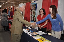 An older man in a tan suit reaches across a table to shake a woman's hand.