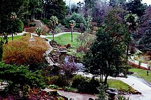 There are tall trees in the background. There is a gazebo below it. There is a walkway leading up to the gazebo. There are trees on both sides of the walkway.