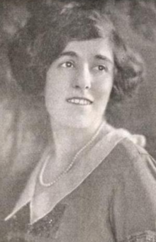 A smiling young white woman, with cropped dark wavy hair, wearing a string of pearls and a dress with a pointed white collar and beaded embellishments