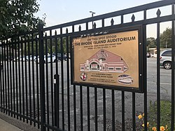 The plaque is hanging on a fence around the current parking lot. It contains the text in the description, along with a diagram of the façade of the former auditorium.
