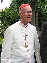 Kardinal Tarcisio Bertone memakai pakaian untuk negara-negara tropika panas (jubah putih dengan perhiasan kain jalur merah dan butang merah).